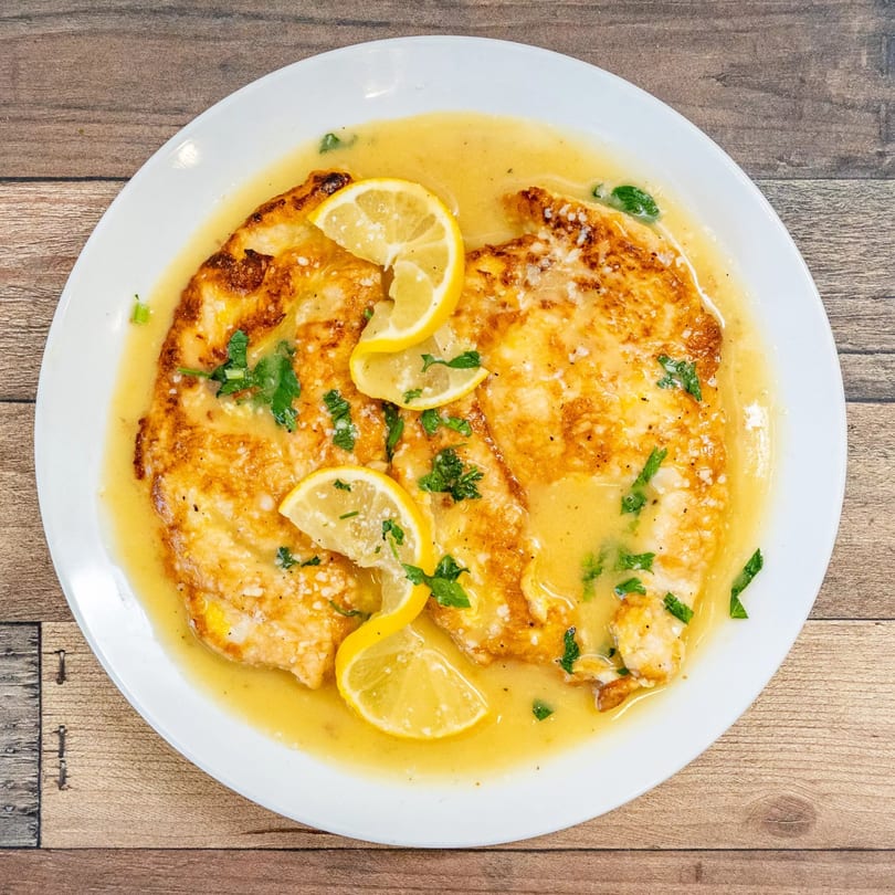 Chicken Francese with lemon slices and parsley in a rich, yellow sauce on a white plate.