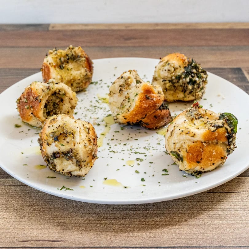 Six golden-brown garlic knots, seasoned with herbs and oil, arranged on a white plate.