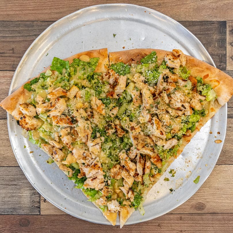 Two slices of Pizza Chop's chicken Caesar pizza with lettuce, chicken, and parmesan on a metal tray.